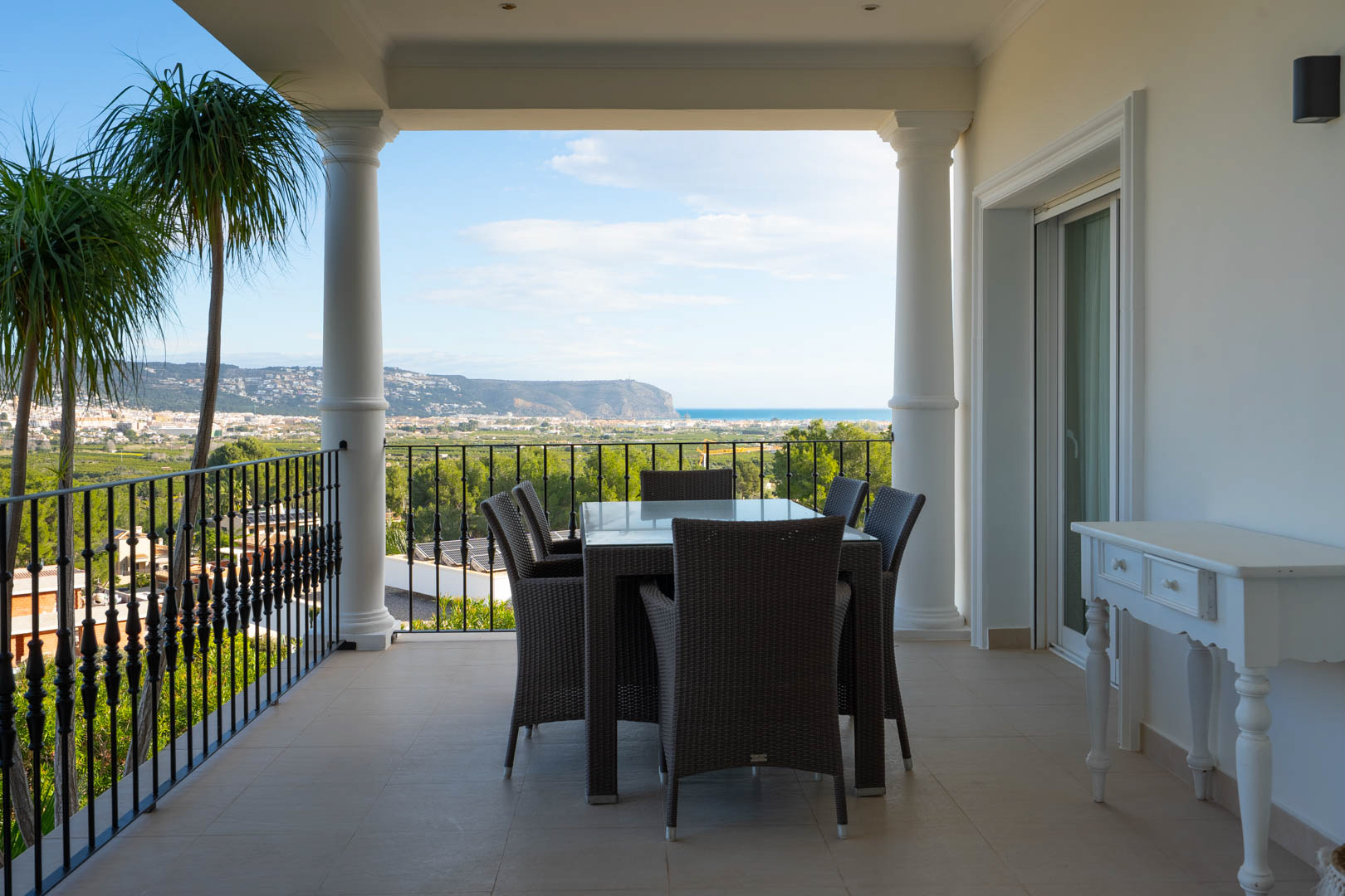 Overdekt terras met klassieke zuilen en smeedijzeren balustrade, voorzien van een eethoek en panoramisch uitzicht op de Middellandse Zee en de vallei.