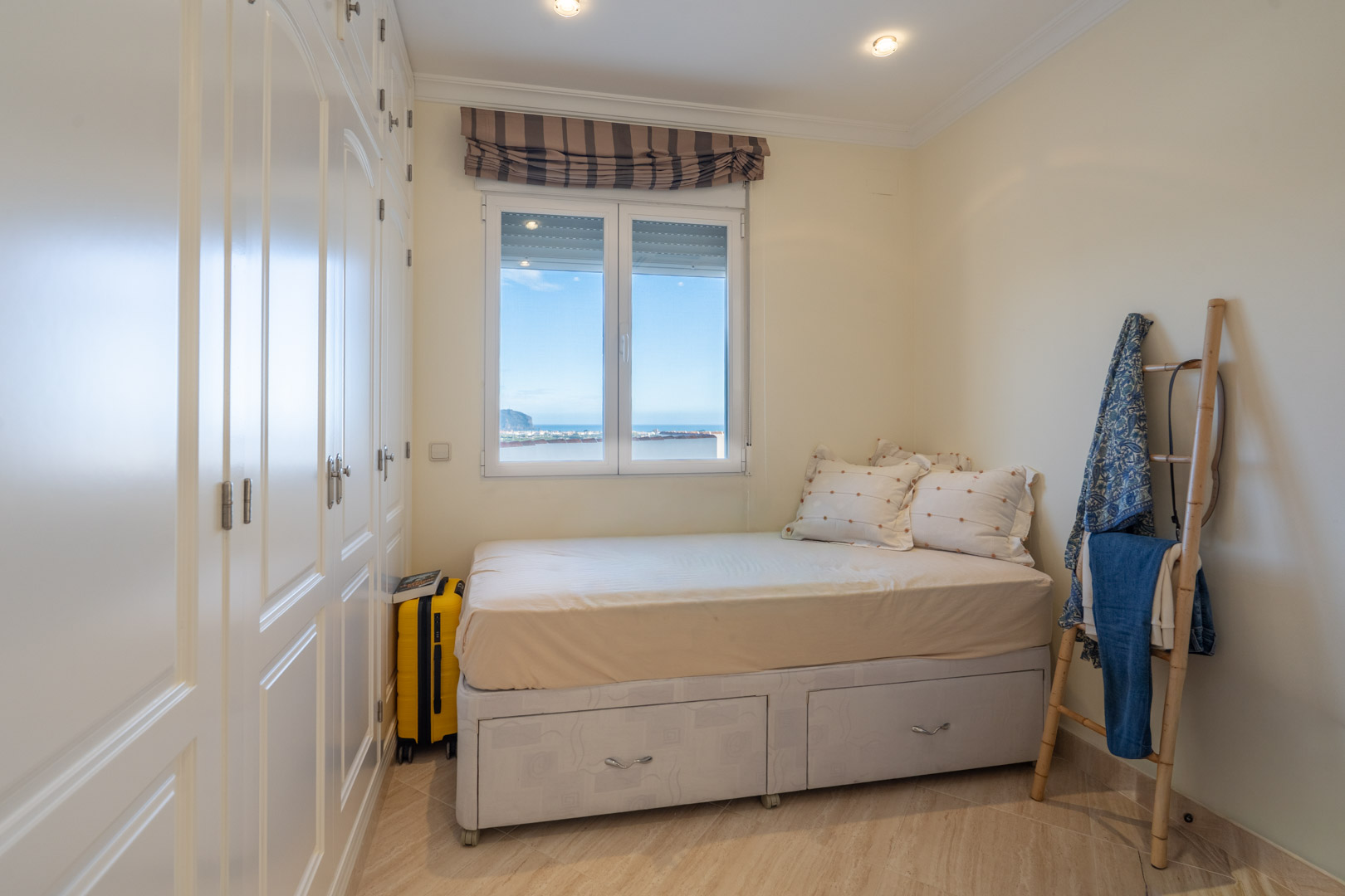 Slaapkamer met witte inbouwkasten, bed met opbergladen en een raam met panoramisch uitzicht over de Middellandse Zee.