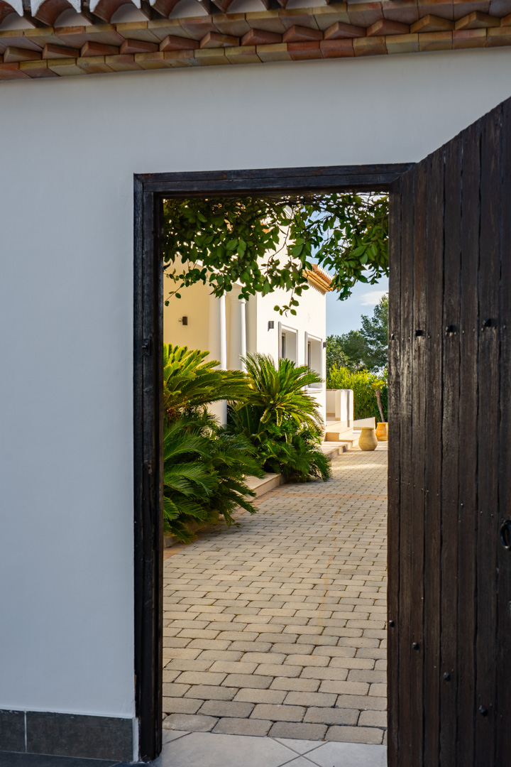 Zicht door een donkere houten deur op het geplaveide pad, omgeven door mediterrane beplanting en de witte gevel van de villa.