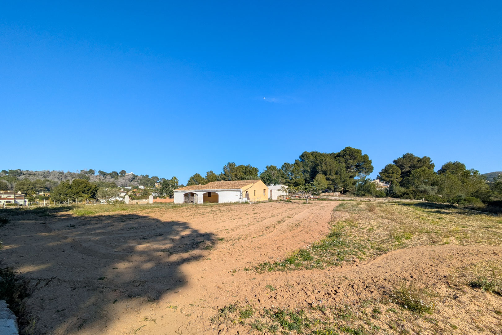 Buitenaanzicht van de traditionele finca met boogterras, gelegen op een groot vlak rustiek perceel omgeven door pijnbomen en natuurlijke landschappen in Jávea.