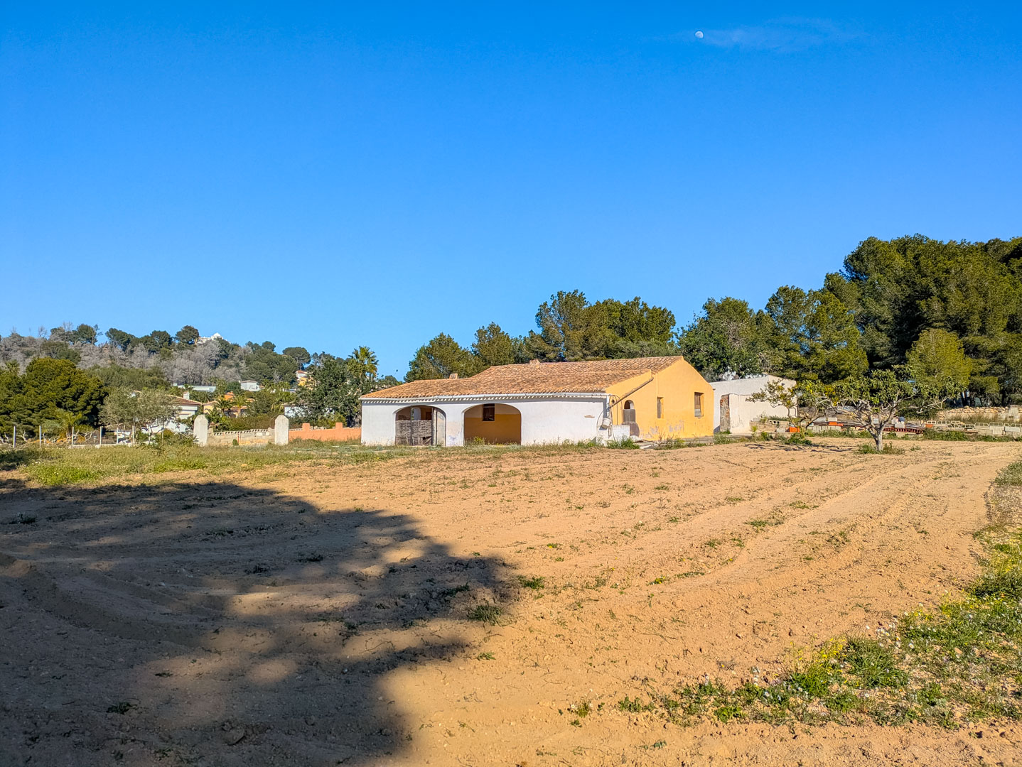 Buitenaanzicht van de traditionele finca met een overdekt terras met bogen, een pannendak en een uitgestrekt vlak rustiek perceel omringd door pijnbomen.