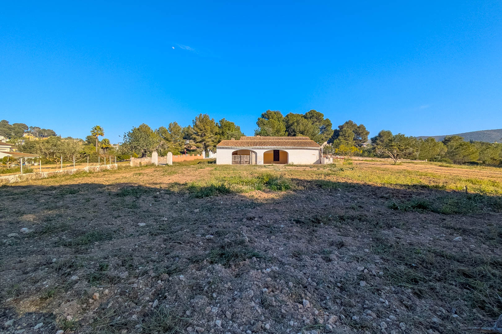 Buitenaanzicht van de traditionele finca met een overdekt terras met bogen en een pannendak, gelegen op een groot vlak perceel omgeven door pijnbomen.