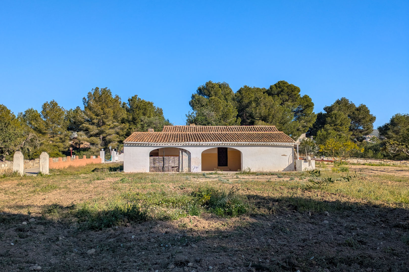 Buitenaanzicht van de traditionele finca met witte gevel, bogenportiek en pannendak, gelegen op een rustiek perceel omgeven door pijnbomen.