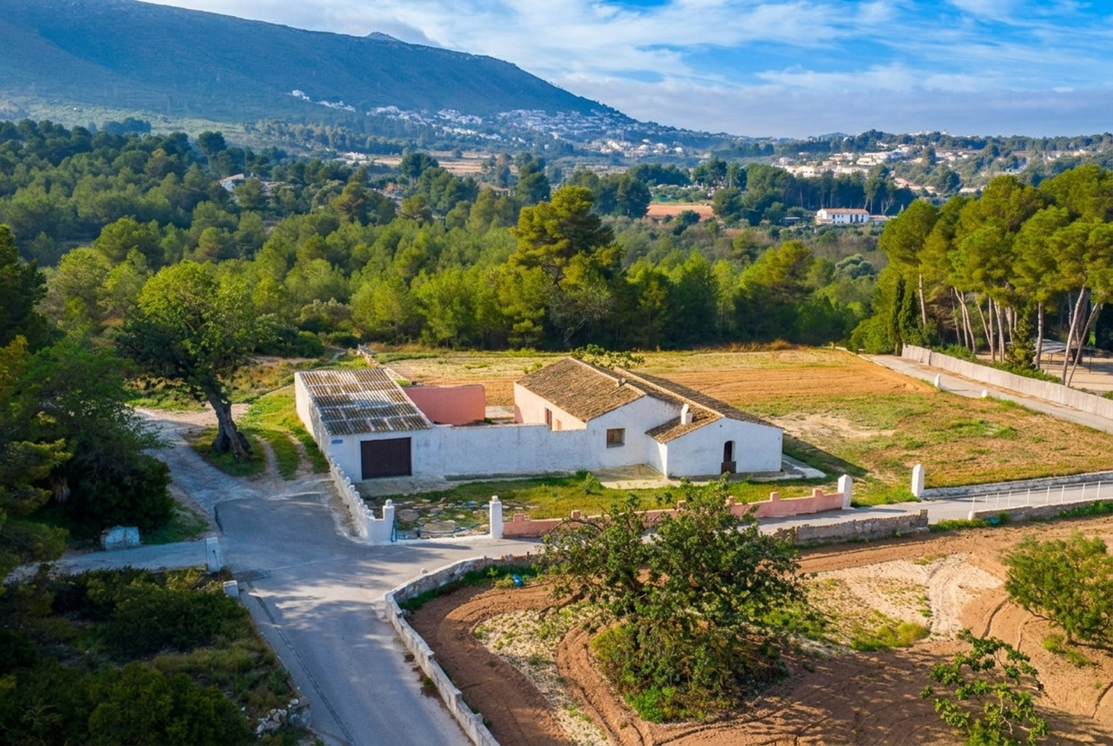 Luchtfoto van de traditionele witgekalkte finca met pannendaken, gelegen op een vlak rustiek perceel omgeven door dennenbossen en uitzicht op de bergen.
