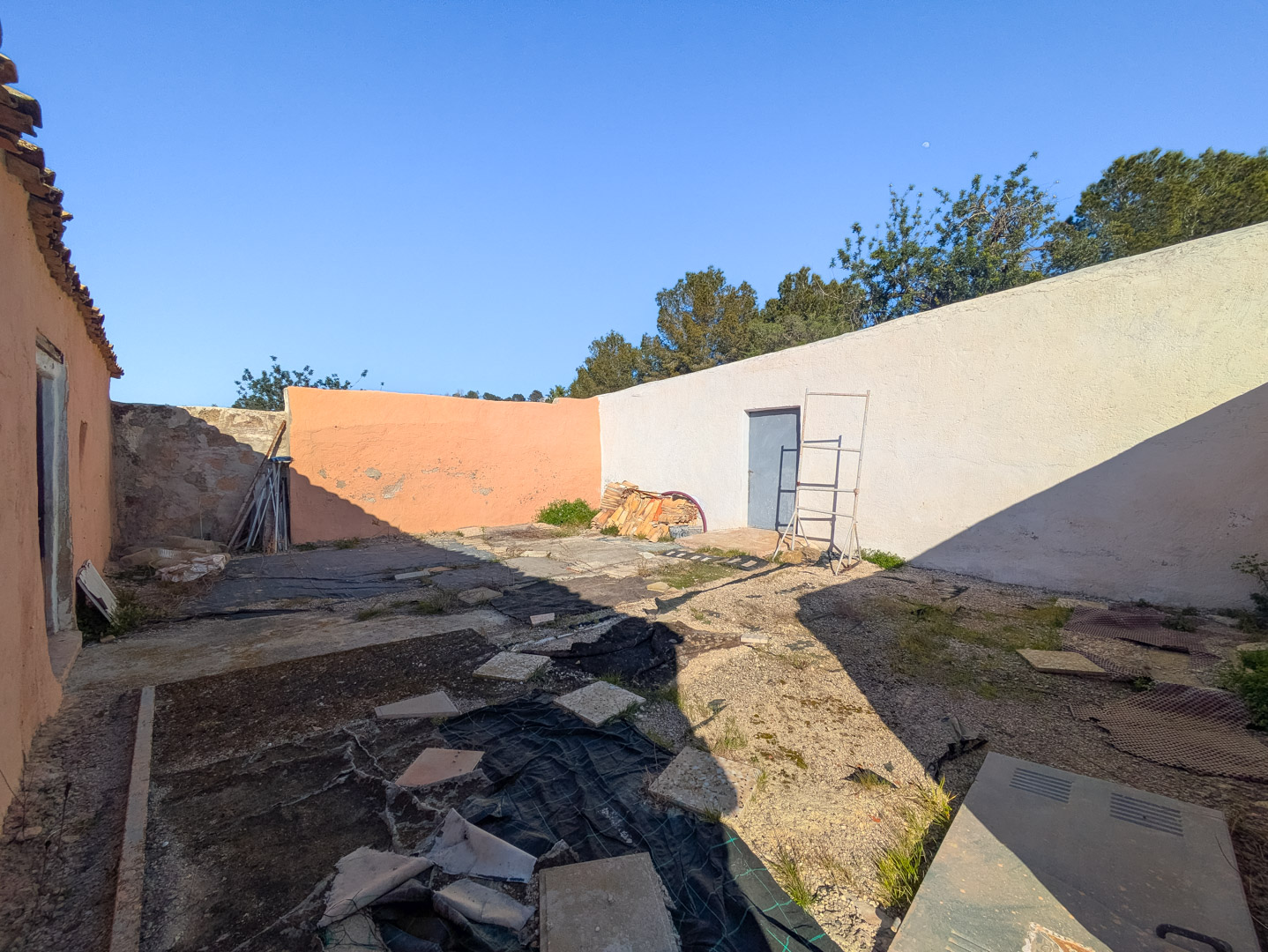 Omsloten binnenplaats met hoge muren, een blauwe deur en een ruwe ondergrond met beschermfolie, klaar voor een volledige renovatie.