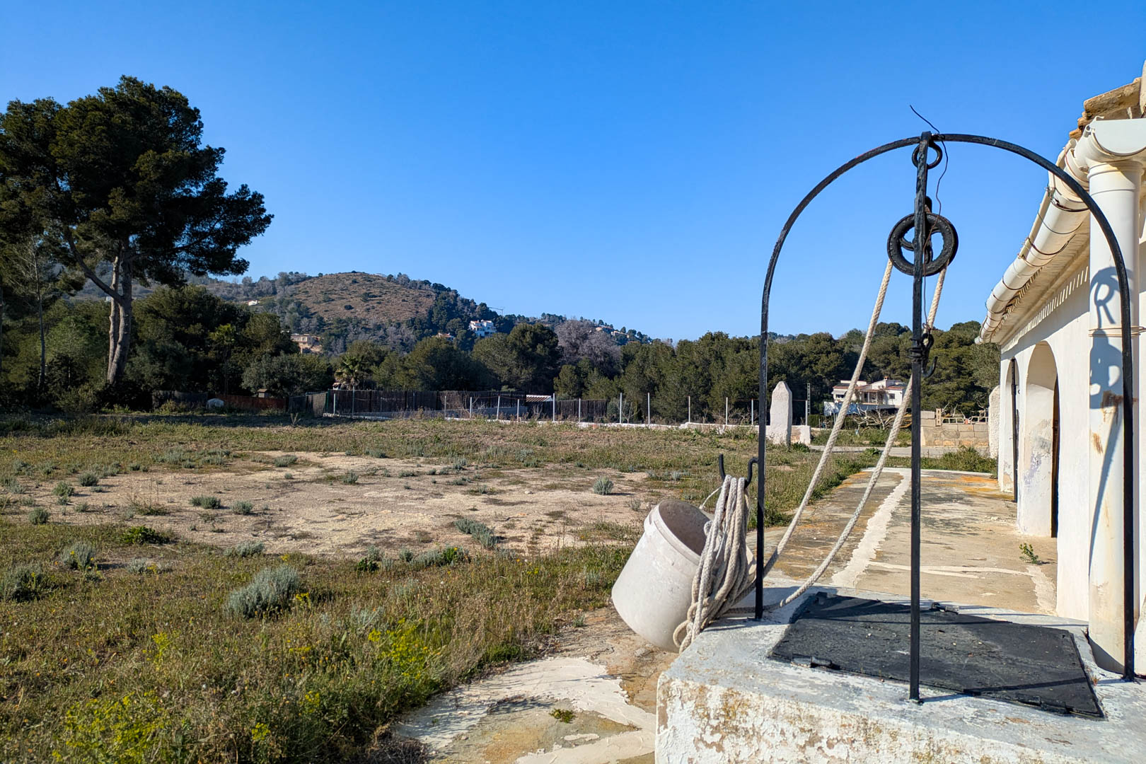 Traditionele waterput met ijzeren boog en katrol op het grote rustieke perceel, met vrij uitzicht op de omliggende heuvels en de natuur van Jávea.