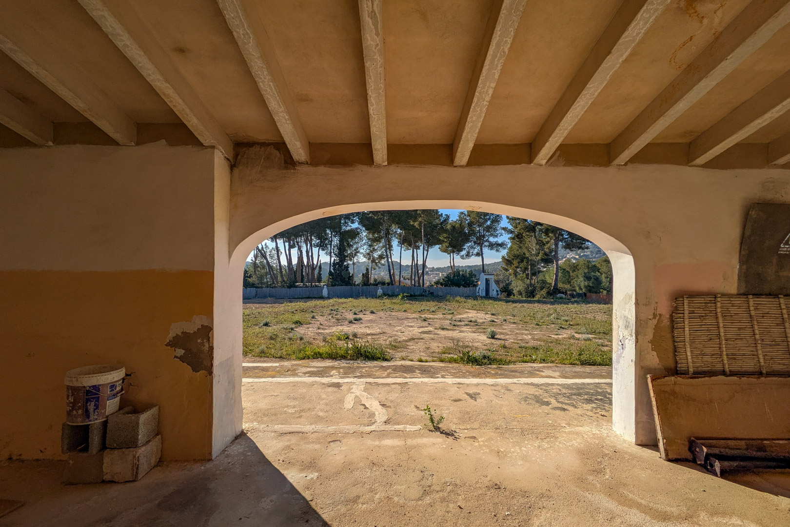 Uitzicht vanuit een gewelfde veranda met balkenplafond op het uitgestrekte rustieke perceel en een rij volwassen pijnbomen op de achtergrond.