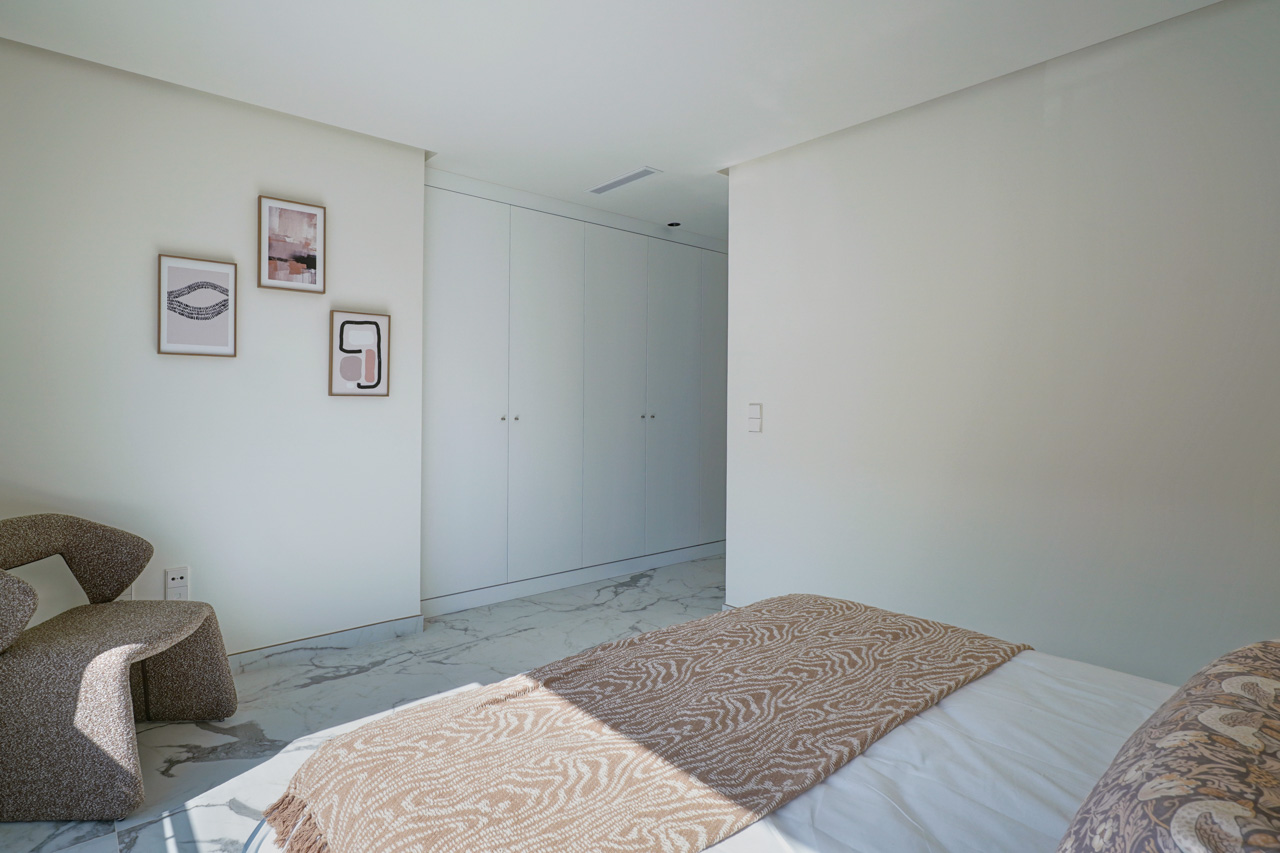 Lichte slaapkamer met kamerhoge witte inbouwkasten, marmerlook vloertegels en een moderne fauteuil met minimalistische wanddecoratie.