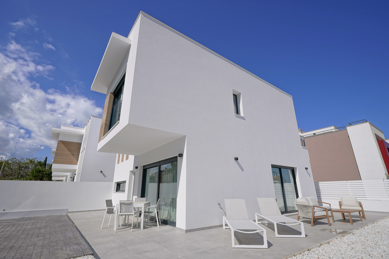 Modern exterieur met witte geometrische volumes, een betegeld terras voorzien van tuinmeubilair en ligbedden, en een eigen geplaveide parkeerplaats.
