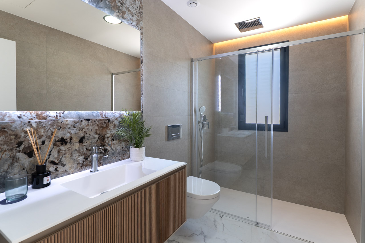 Moderne badkamer met houten lamellenmeubel, marmeren achterwand en inloopdouche met glazen wand en indirecte plafondverlichting.