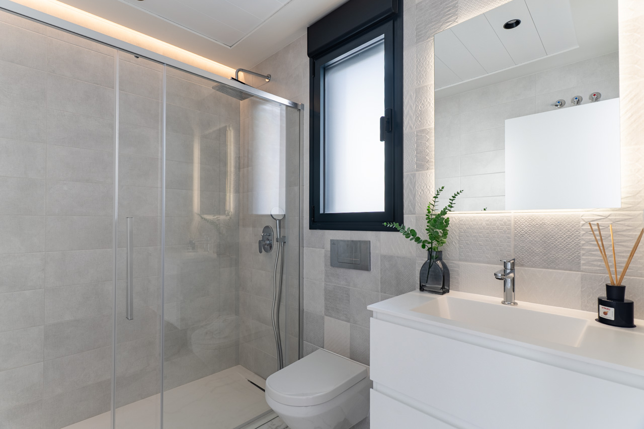 Moderne badkamer met inloopdouche, glazen wand, getextureerde grijze tegels, wit wastafelmeubel en een verlichte spiegel met indirecte LED-verlichting.