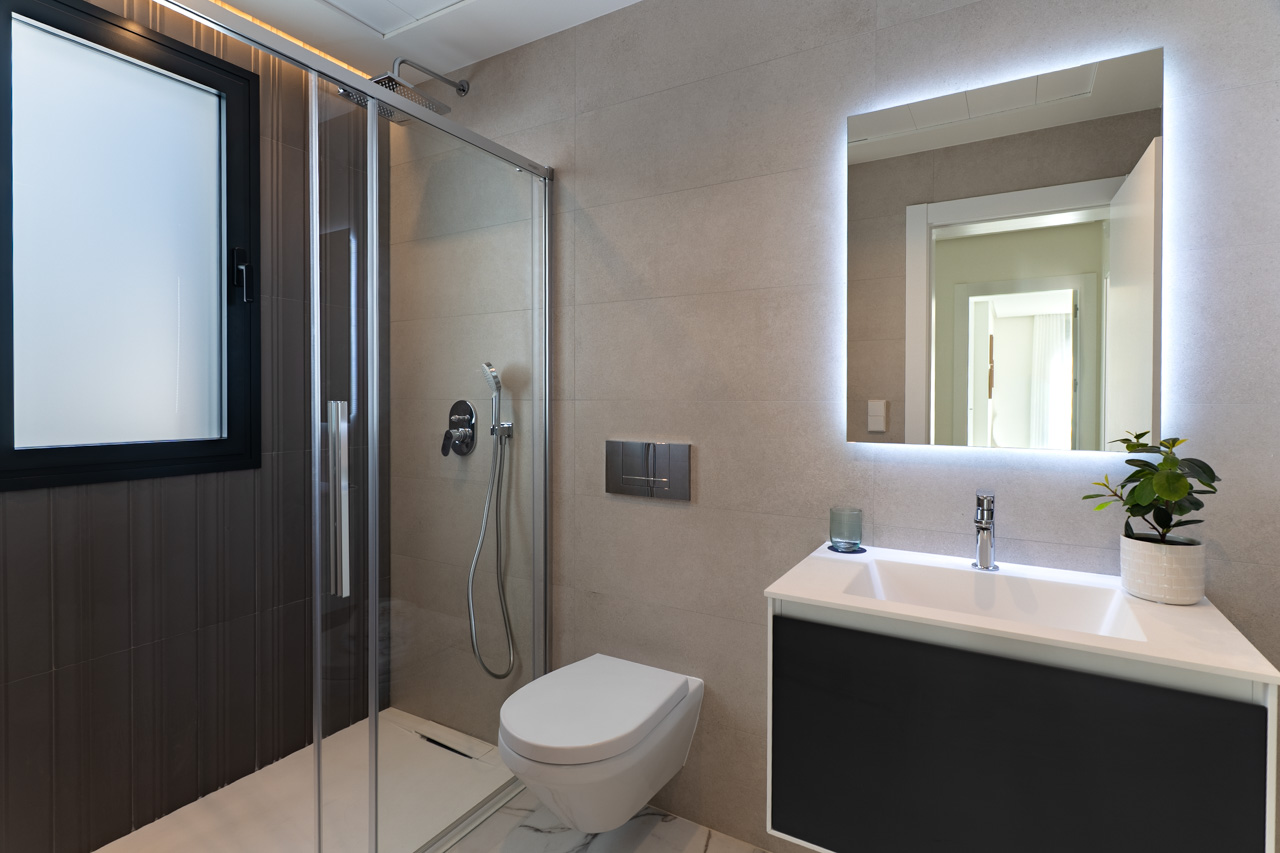 Moderne badkamer met inloopdouche, glazen wand, hangend toilet en een wastafelmeubel met indirect verlichte spiegel tegen een achtergrond van neutrale tegels.