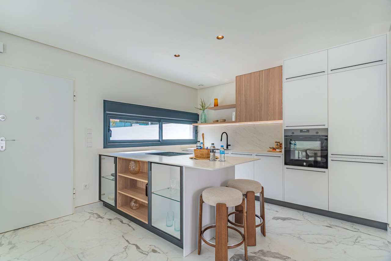 Moderne open keuken met kookeiland, witte kasten en houten accenten. Voorzien van inbouwapparatuur en een vloer van porselein met marmerlook.