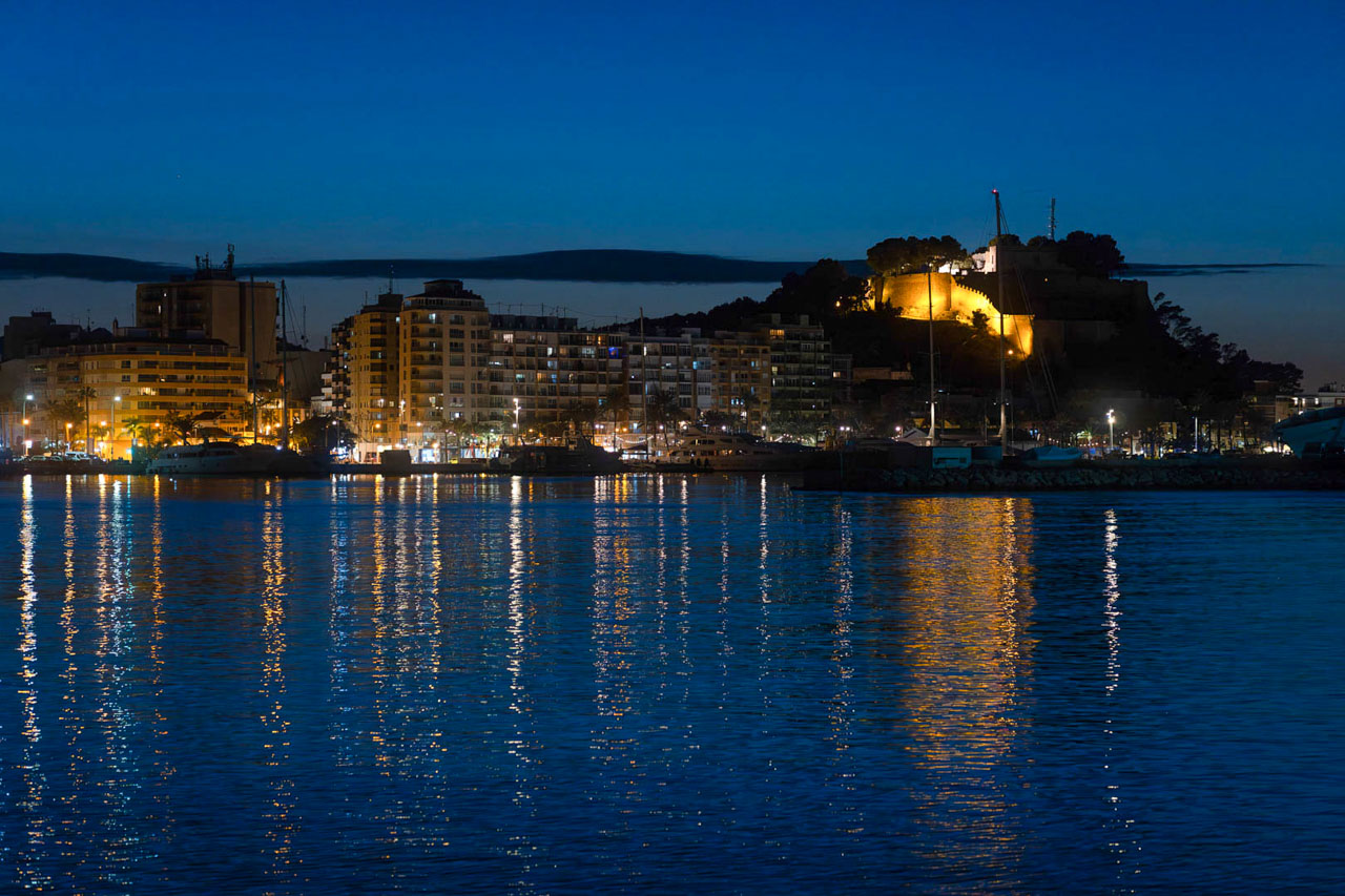 Nachtzicht op de haven van Dénia met het verlichte kasteel op de heuvel en de weerspiegeling van de stadslichten op het water van de Middellandse Zee.