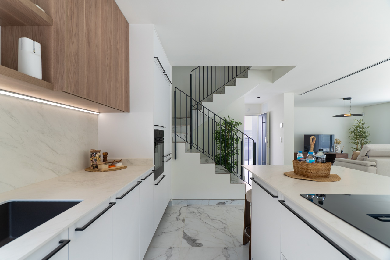Open keuken met witte kasten, marmerlook werkbladen en houten bovenkasten met LED-verlichting. Op de achtergrond zijn de moderne trap en de woonkamer zichtbaar.