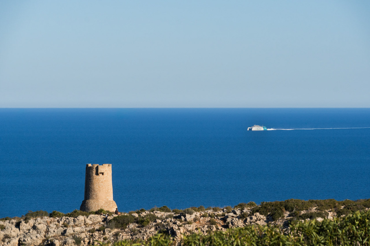 Panoramisch uitzicht op de kustlijn van Las Rotas in Dénia, met de historische Torre del Gerro-wachttoren en een Balearia-veerboot op de Middellandse Zee.