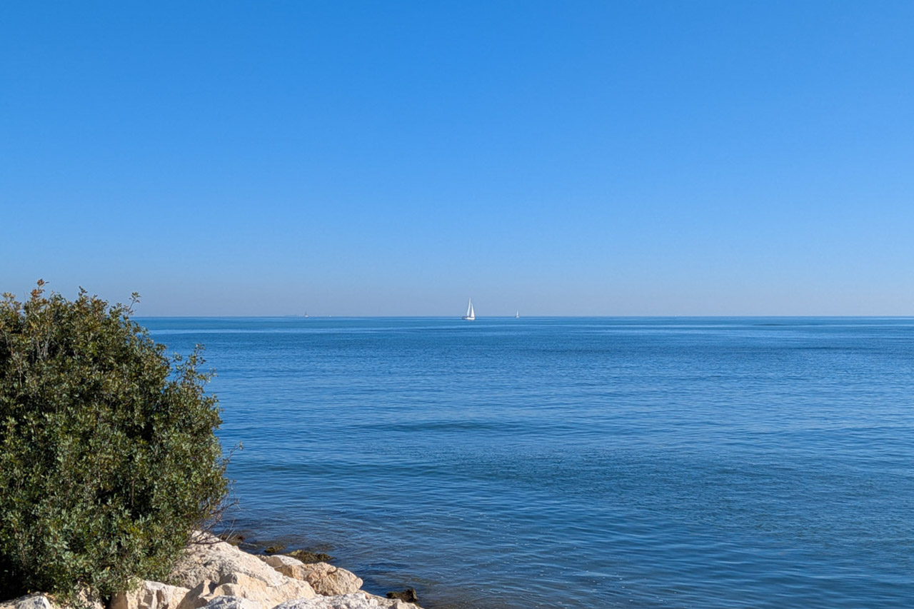Panoramisch uitzicht op de Middellandse Zee vanaf de rotsachtige kust van Dénia, met zeilboten aan de horizon onder een helderblauwe hemel.