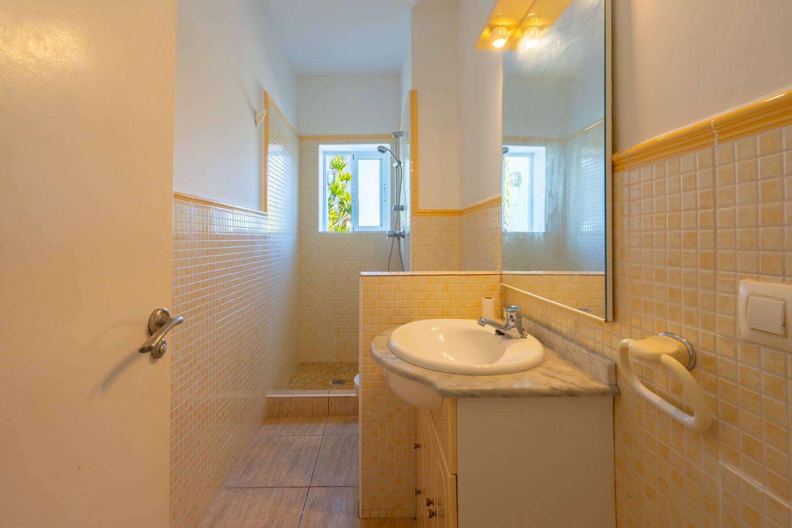 Badkamer met inloopdouche, geel-beige mozaïektegels, een wastafelmeubel met marmeren blad en natuurlijke lichtinval via een raam.