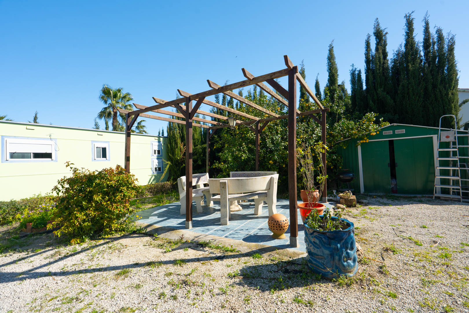 Buitenruimte met houten pergola en stenen banken op een betegeld terras, gesitueerd tussen de berging en het geprefabriceerde gastenverblijf.