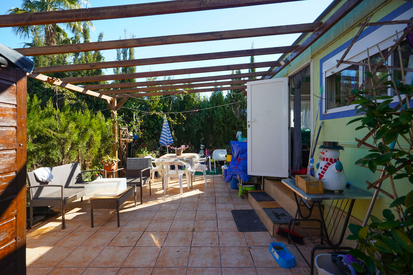 Buitenterras van de geprefabriceerde woning met keramische tegels, houten balkenpergola en een zithoek omgeven door privacybiedende hagen.