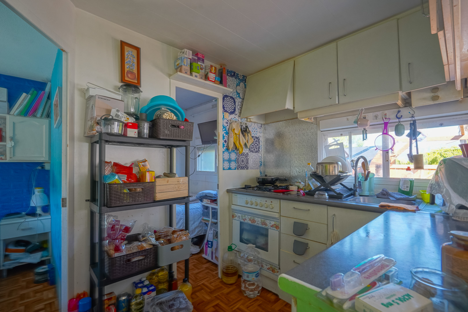 Compacte keuken in het bijgebouw met witte kasten, een decoratieve tegelwand bij het fornuis en open opbergrekken, verlicht door een raam boven de gootsteen.