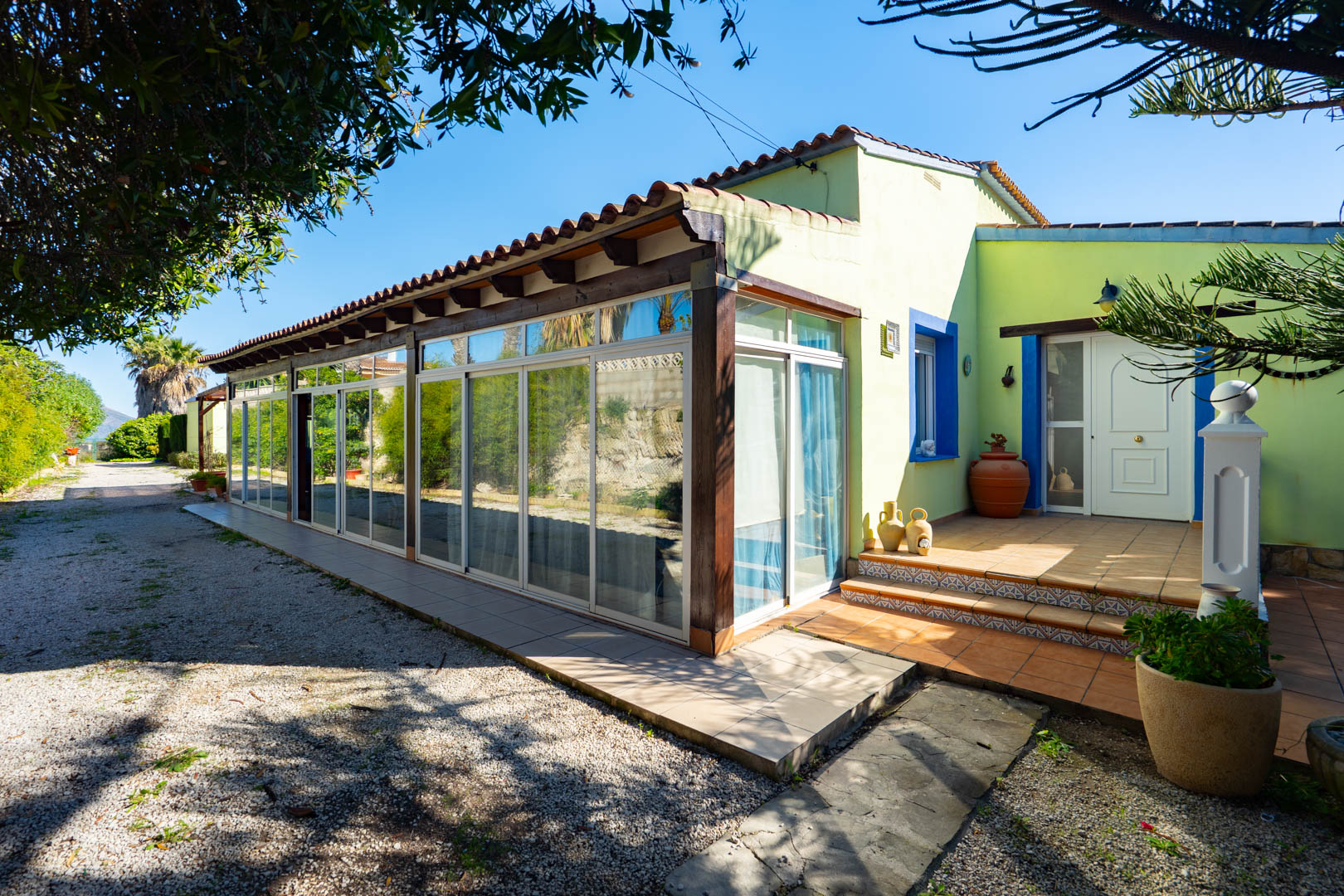 Exterieur van de finca met een glazen naya met houten balken en een betegelde entree met mediterrane blauwe accenten.
