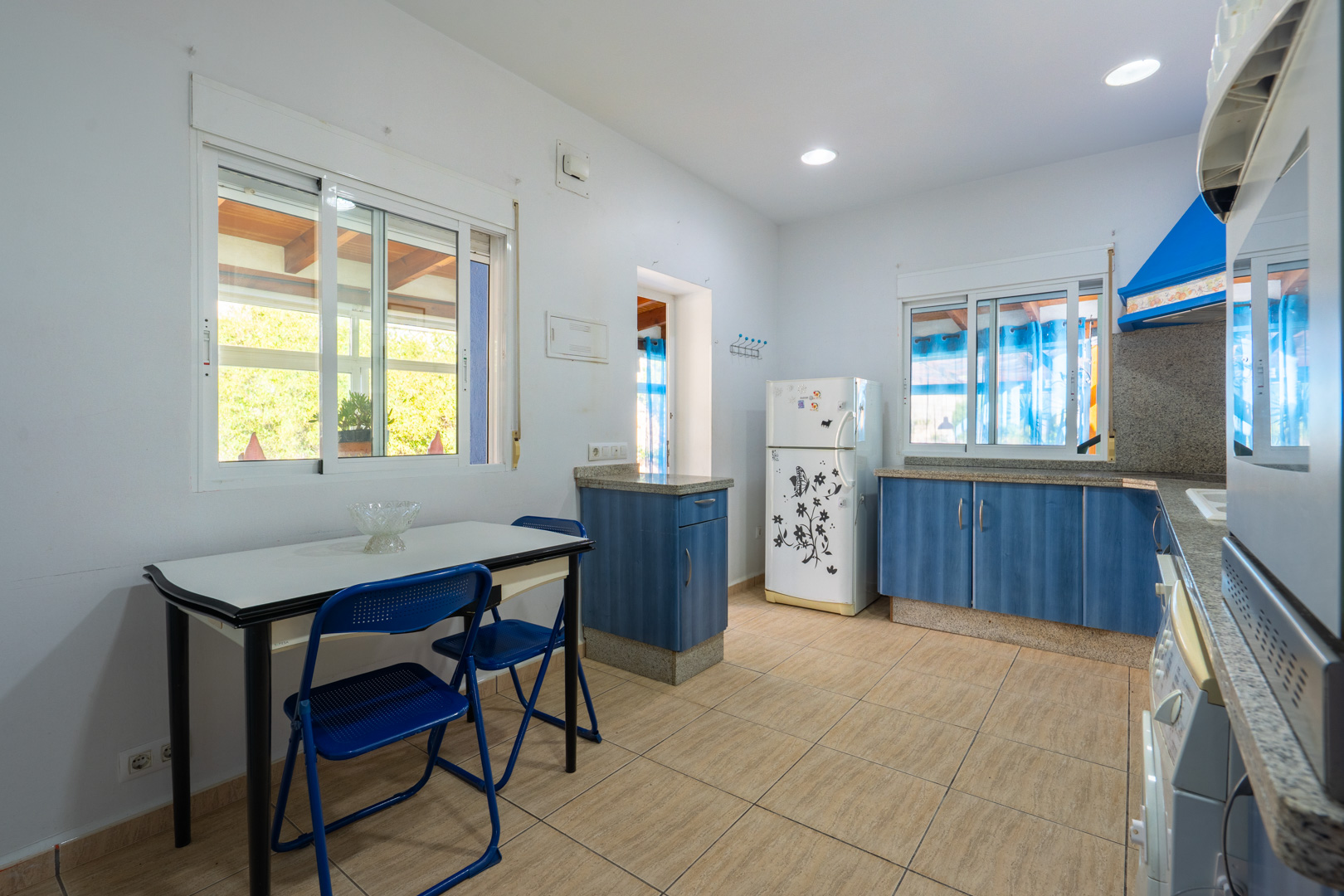 Functionele keuken met blauwe kasten, granieten werkbladen en een eethoek. Voorzien van inbouwspots en ramen die uitkijken op het overdekte terras.