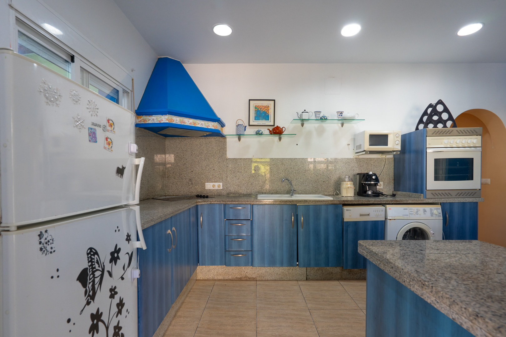 Functionele keuken met blauwe kasten, grijs granieten werkbladen en bijpassende achterwand. Voorzien van een blauwe afzuigkap, inbouwapparatuur en inbouwspots.