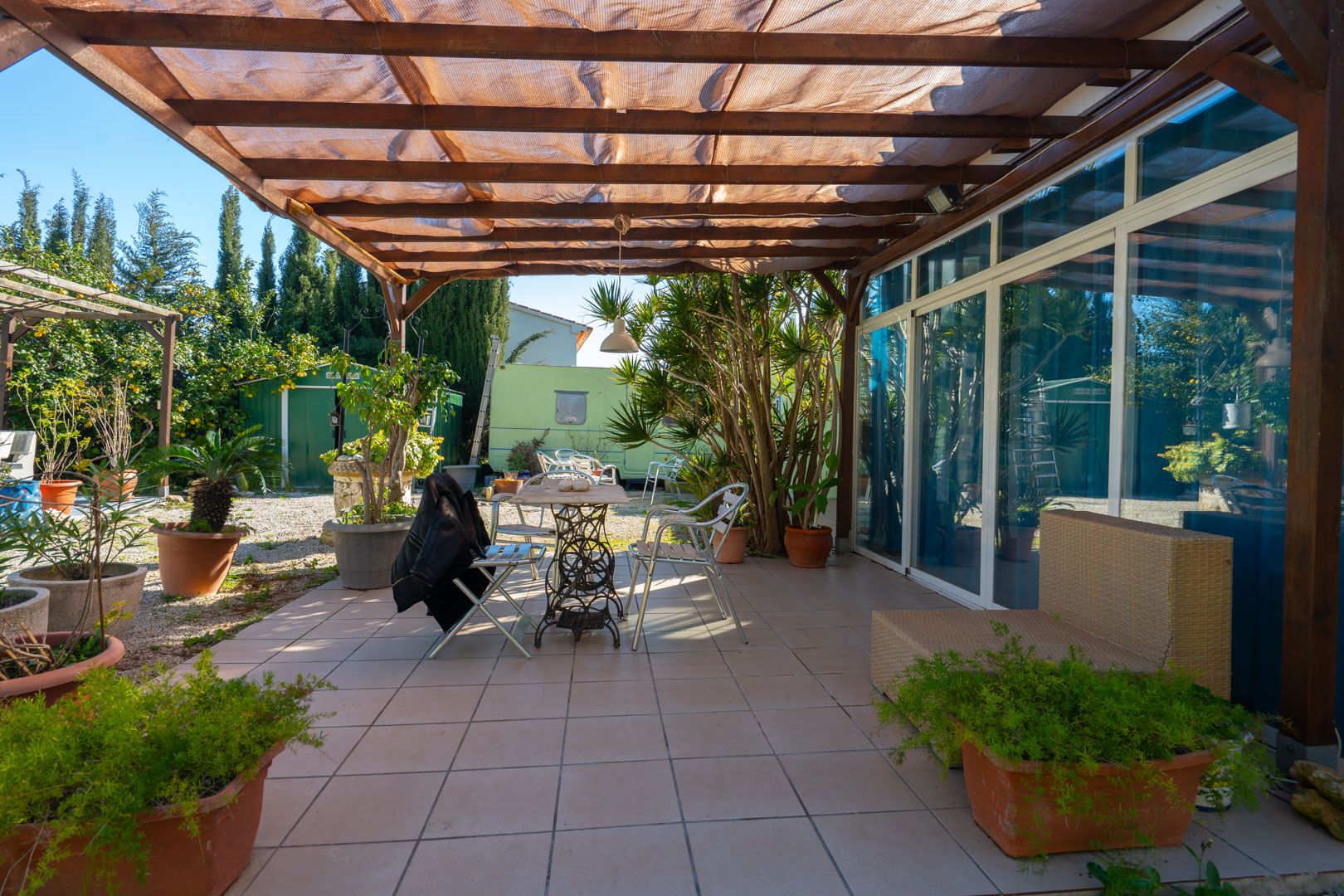 Geplaveid terras met houten pergola en schaduwdoek, voorzien van buitenmeubilair en diverse potplanten grenzend aan de mediterrane tuin.
