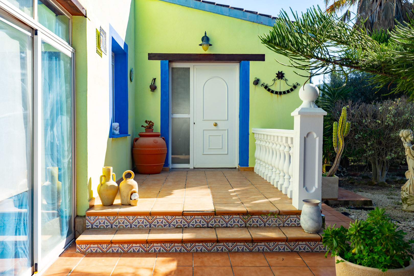 Hoofdingang met een witte deur, blauwe kozijnen en decoratieve tegels op de treden. Het terracotta terras is voorzien van een witte balustrade en mediterrane potten.