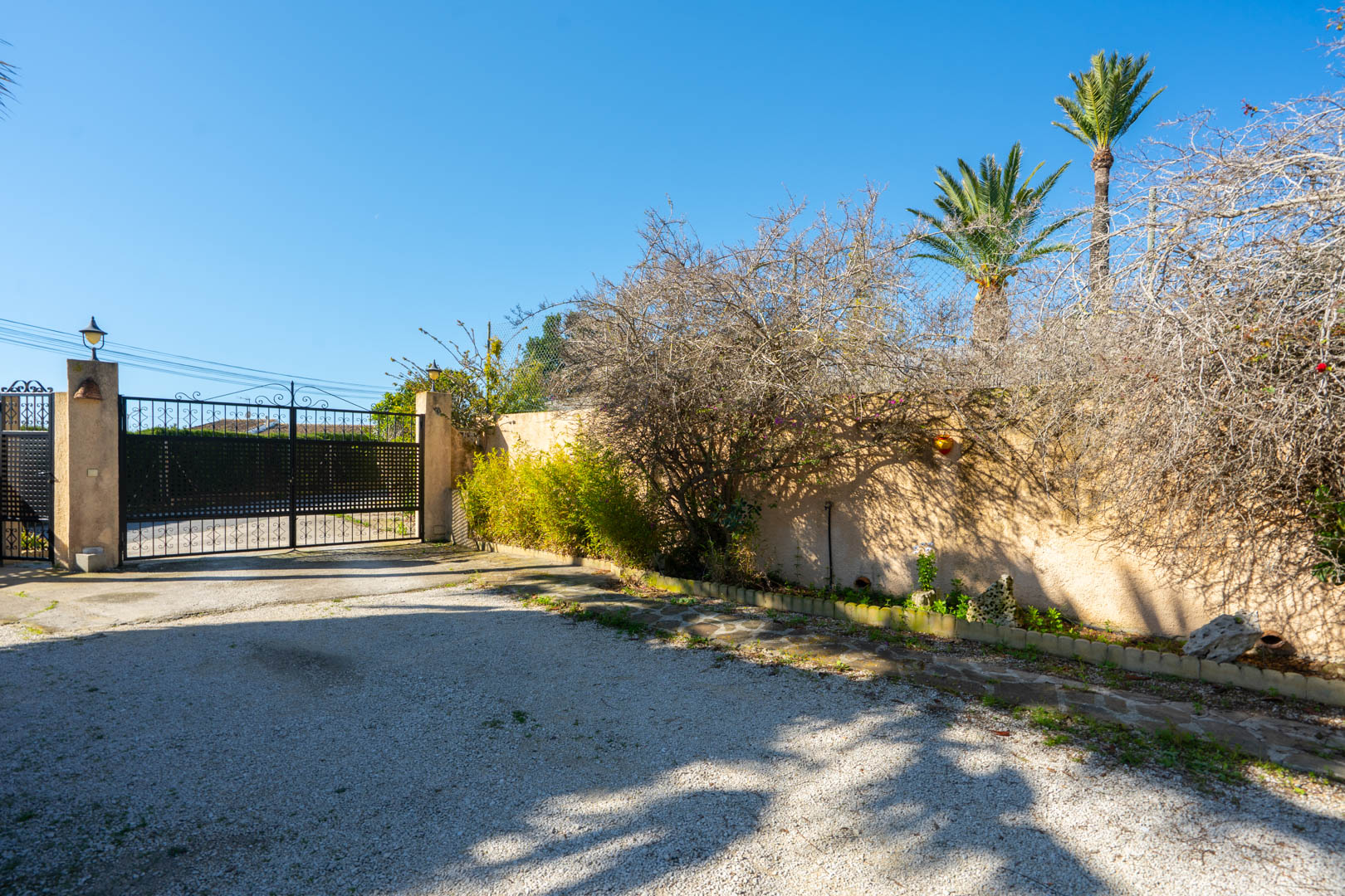 Hoofdingang met een zwart smeedijzeren poort, een oprit van grind en een omheiningsmuur met mediterrane beplanting en palmbomen.