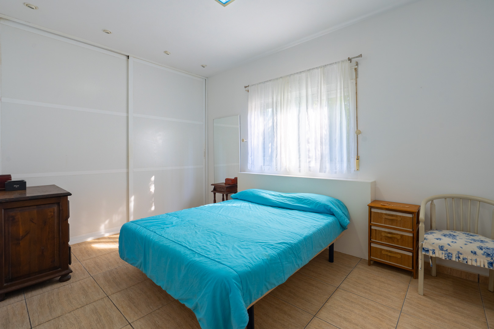 Lichte slaapkamer met witte inbouwkasten, tegelvloer, raam met transparante gordijnen en klassiek houten meubilair.