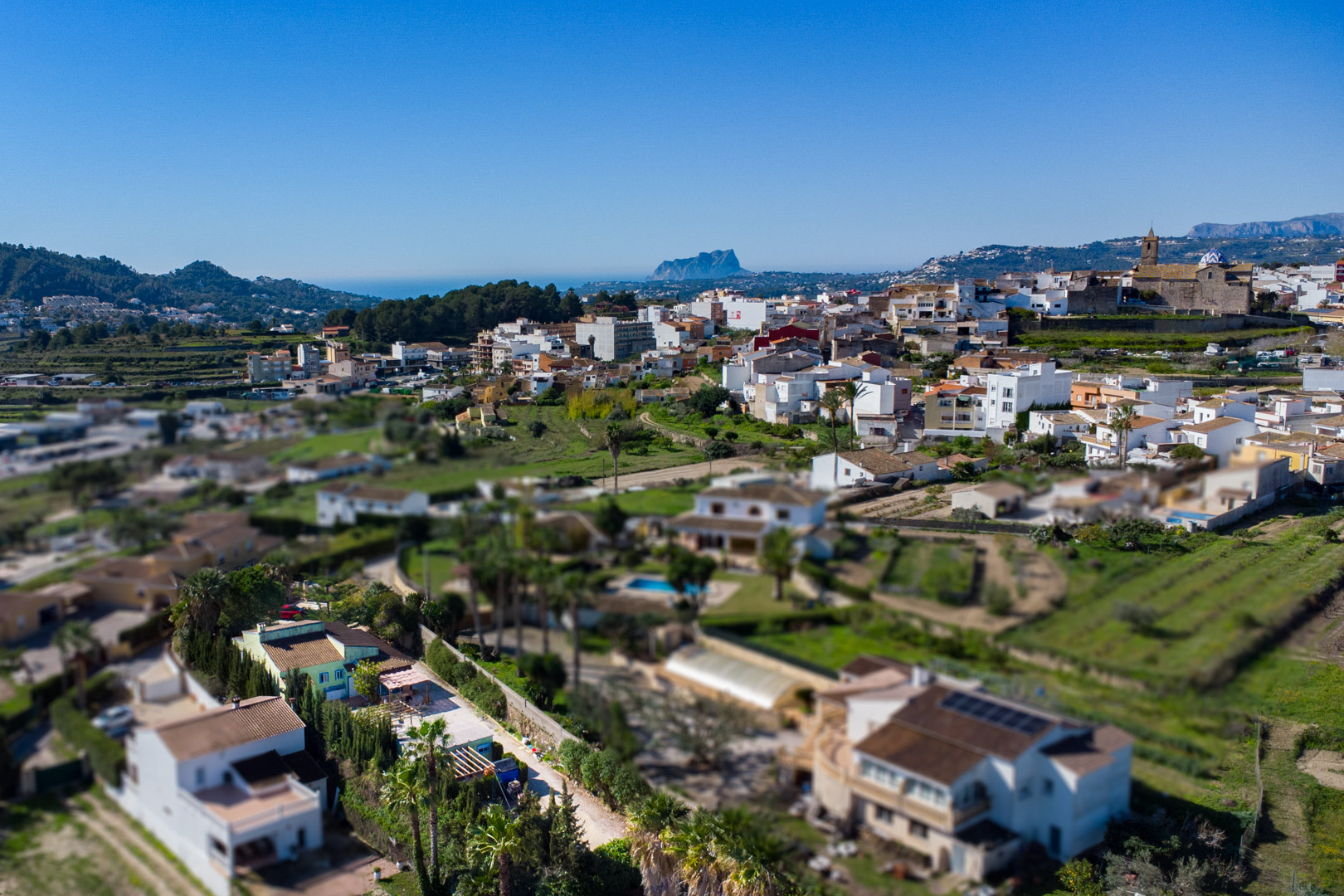Panoramisch luchtfoto van Benitachell met de Peñón de Ifach aan de horizon. Het toont de dorpskern, de parochiekerk en het omliggende mediterrane landschap richting de zee.
