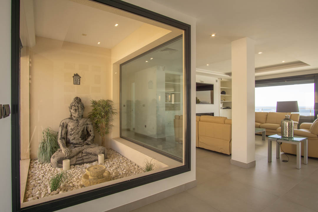 Beglaasde binnentuin in zen-stijl met Boeddhabeeld en witte keien, grenzend aan een ruime woonkamer met inbouwspots en uitzicht op zee.
