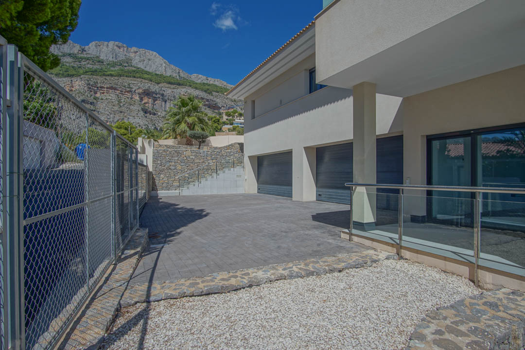 Buiteningang met drievoudige garage, geplaveide oprit, natuurstenen muren en glazen balustrades met uitzicht op de Sierra de Altea.