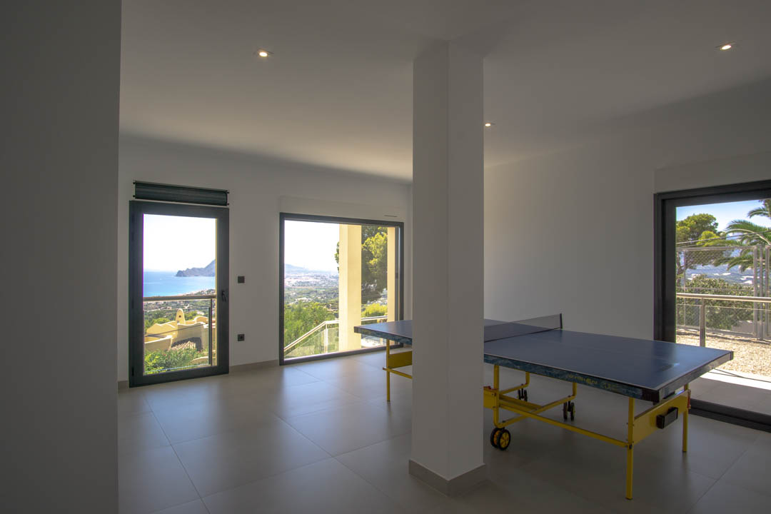 Gameroom met tafeltennistafel, grijze tegelvloer en grote ramen met panoramisch uitzicht over de kust van Altea en de Middellandse Zee.
