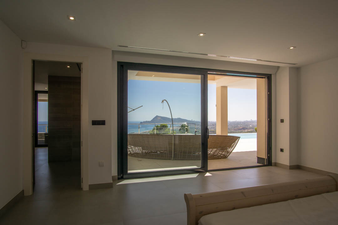 Hoofdslaapkamer met grote glazen schuifdeuren naar het terras, panoramisch uitzicht op zee, inbouwspots en een minimalistische tegelvloer.