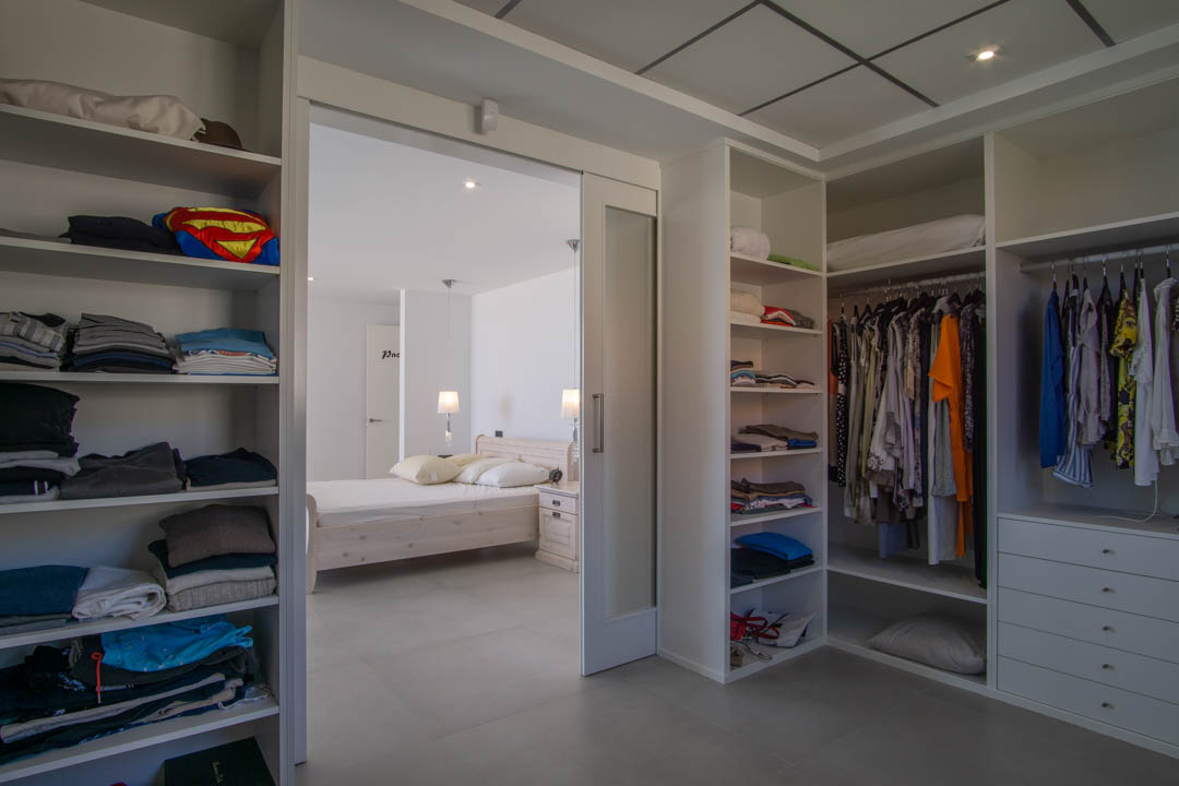 Inloopkast van 12m2 met witte modulaire kasten, lades en een glazen schuifdeur die toegang biedt tot de hoofdslaapkamer met inbouwspots.