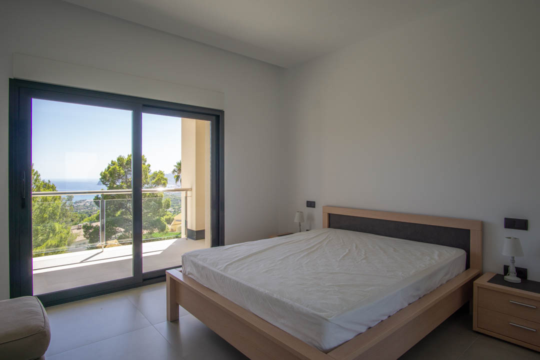 Lichte slaapkamer met een licht houten bedframe, directe toegang tot een privébalkon en vrij uitzicht op de Middellandse Zeekust en pijnbomen.