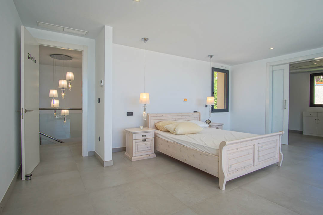 Lichte slaapkamer met licht houten bedframe, bijpassende nachtkastjes en hanglampen. Grootformaat tegelvloer en toegang tot de gang met een glazen balustrade.