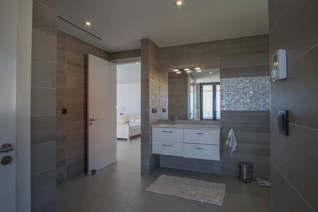 Moderne badkamer met grijze wandtegels, wit zwevend wastafelmeubel en mozaïekdetails. De open verbinding met de slaapkamer biedt zicht op de verre horizon.