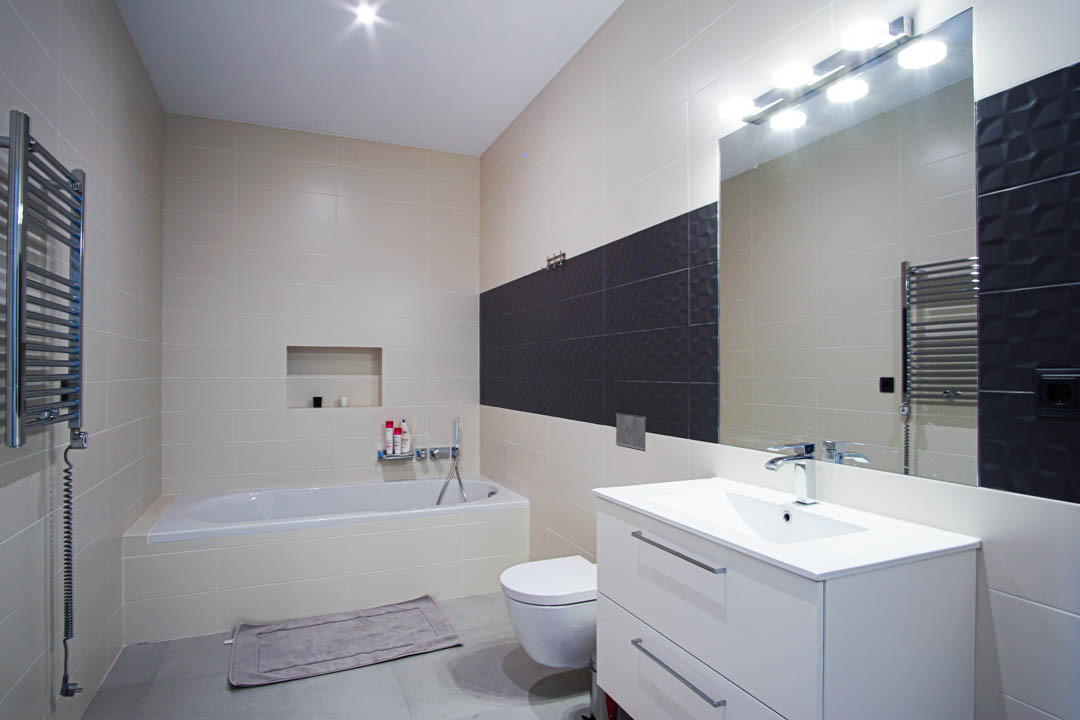 Moderne badkamer met ingebouwd ligbad, wit wastafelmeubel, hangend toilet en beige wandtegels met een donkergrijze, getextureerde accentstrip.