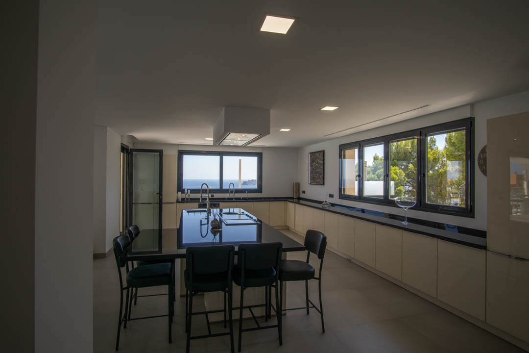 Moderne keuken met een groot kookeiland met zwart werkblad, hangende afzuigkap en kamerhoge ramen met uitzicht op de Middellandse Zee en de omliggende pijnbomen.