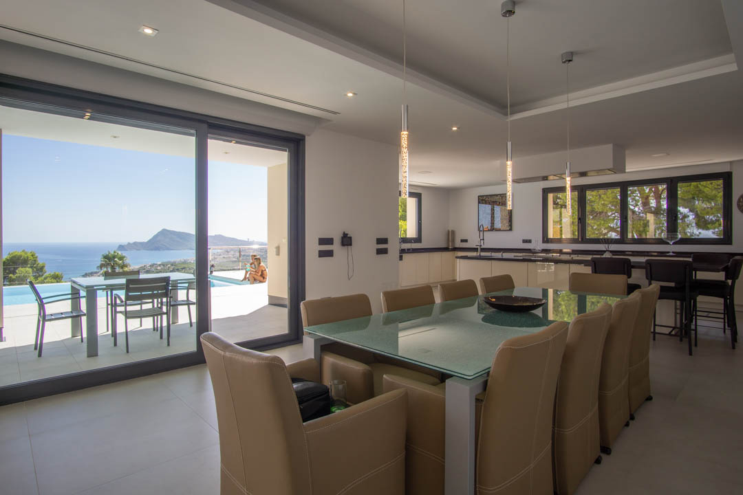 Open eetkamer met glazen tafel, moderne keuken en schuifpui naar het terras met panoramisch uitzicht over de baai van Altea en het zwembad.