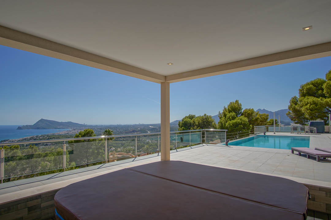 Overdekt terras met jacuzzi, buitenzwembad en panoramisch uitzicht over de baai van Altea en de skyline van Benidorm, afgewerkt met glazen en stalen balustrades.