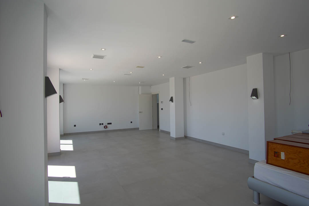 Ruime open kamer met grijze tegelvloer, witte muren, inbouwspots in het plafond en moderne wandverlichting.