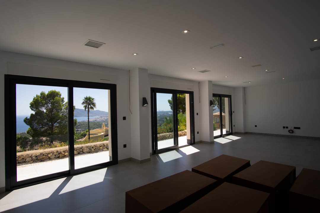Ruime open woonkamer met grote schuifpuien met zwart frame, inbouwspots in het plafond en panoramisch uitzicht over de Middellandse Zee en de kustlijn van Altea.