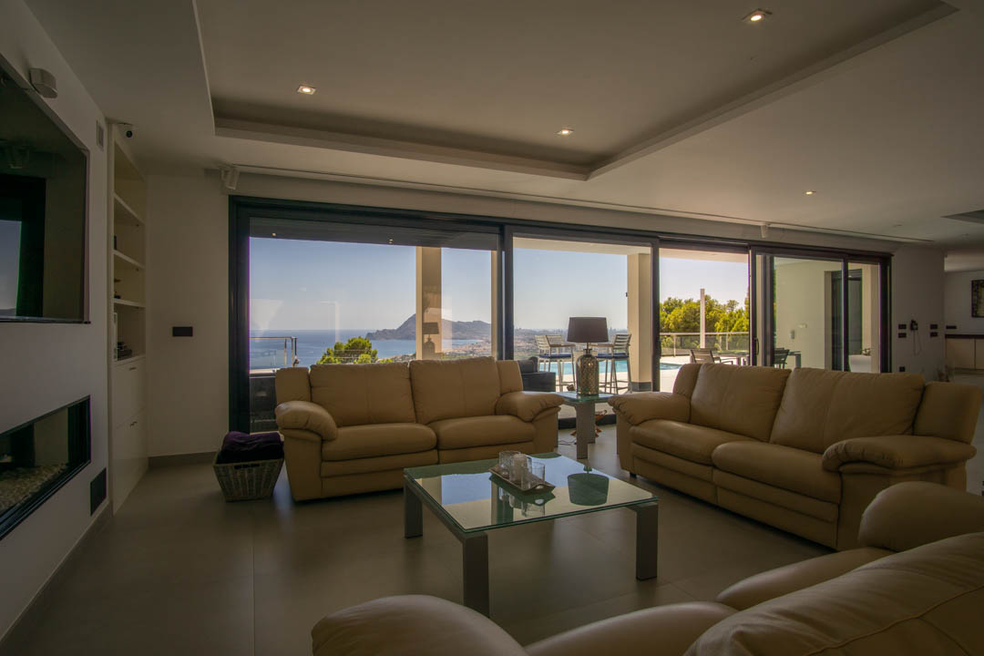 Ruime woonkamer met lederen banken, inbouwverlichting en kamerhoge glazen schuifdeuren met panoramisch uitzicht op de zee en de baai van Altea.