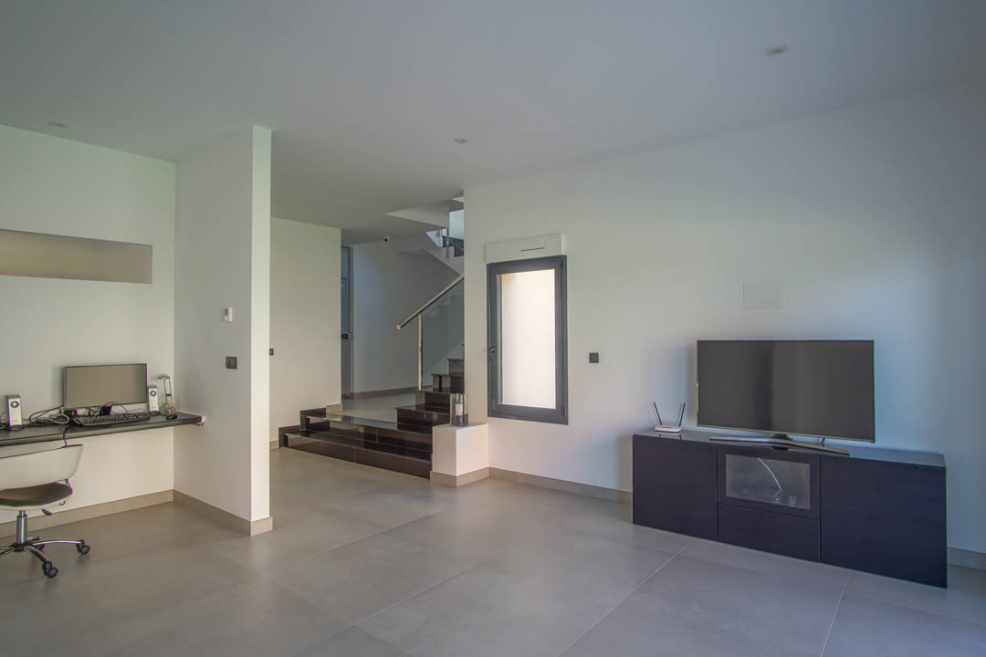 Tweede woonkamer met geïntegreerde werkplek, grijze tegelvloer en een moderne trap met glazen balustrade die naar de bovenverdieping leidt.