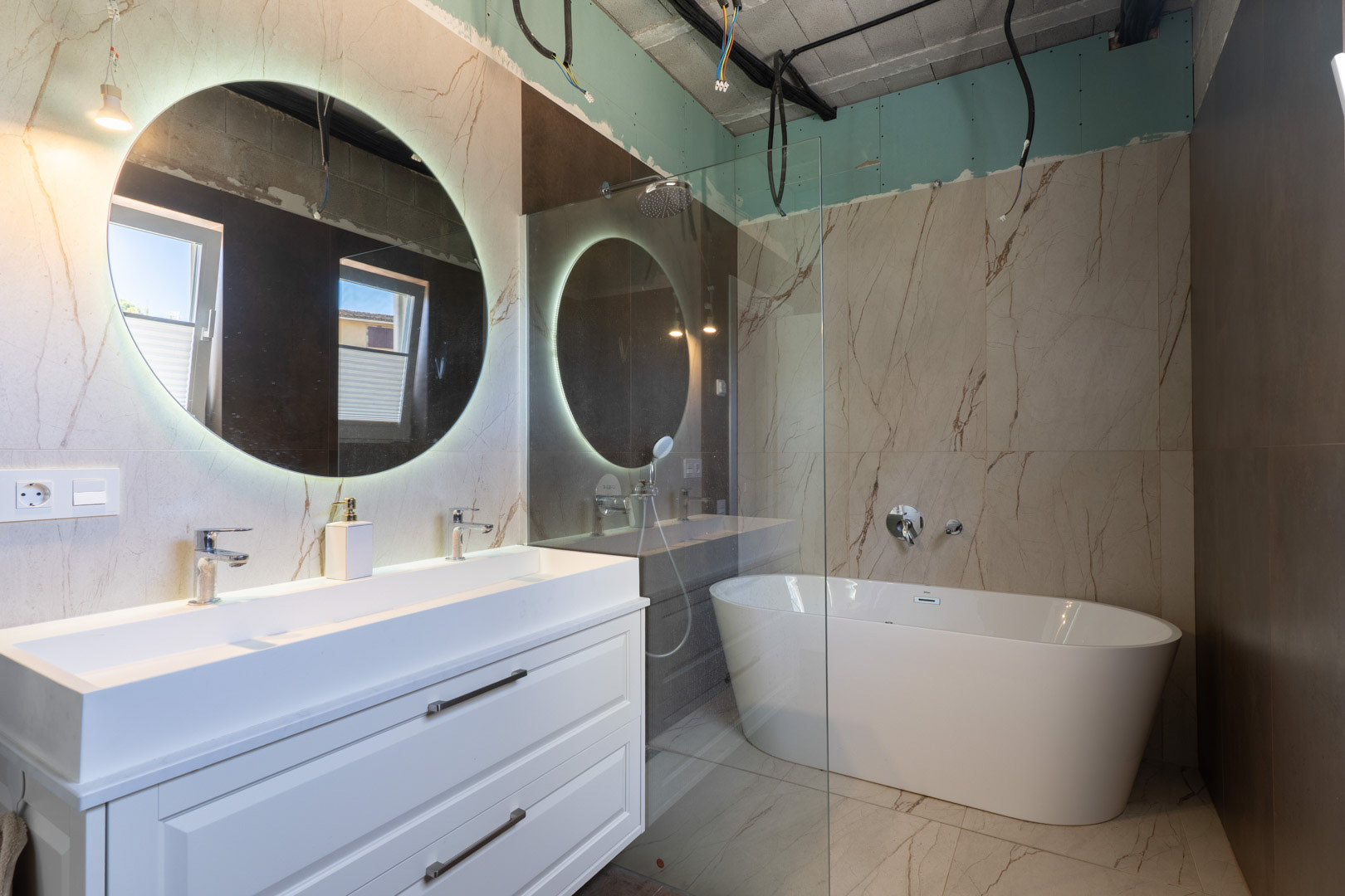 Badkamer in afbouw met dubbele wastafel, verlichte ronde spiegel en vrijstaand ligbad tegen wanden met marmerlook porseleinen tegels.
